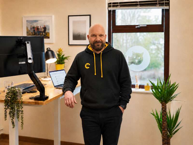 MarkStandingInStudio Bald man in black and yellow hoodie standing by a standing desk
